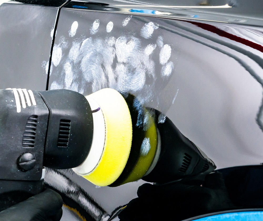 A polishing tool with a yellow pad is being used to buff the surface of a black car, showcasing the Paint Correction in New Orleans process, with visible polishing compound residue.