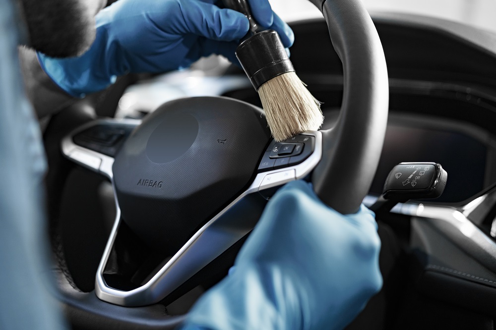 A person wearing blue gloves cleans a car steering wheel with a brush, carefully focusing on the control buttons and central area—demonstrating meticulous Car Detailing in Kenner.