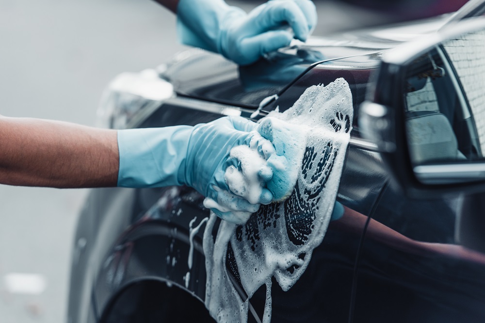 A person wearing blue gloves washes a black car with a soapy sponge, focusing on the exterior surface—a perfect example of expert Auto Detailing Covington services.