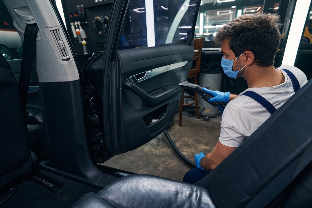 A worker in overalls and a mask cleans the interior of a car with the door open in a garage, showcasing professional Auto Detailing Covington services.